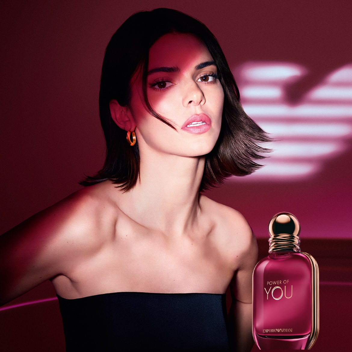 Una donna con un top nero senza spalline e orecchini dorati mette in mostra il profumo Power of You di Emporio Armani, il cui flacone è di un elegante color rosa.