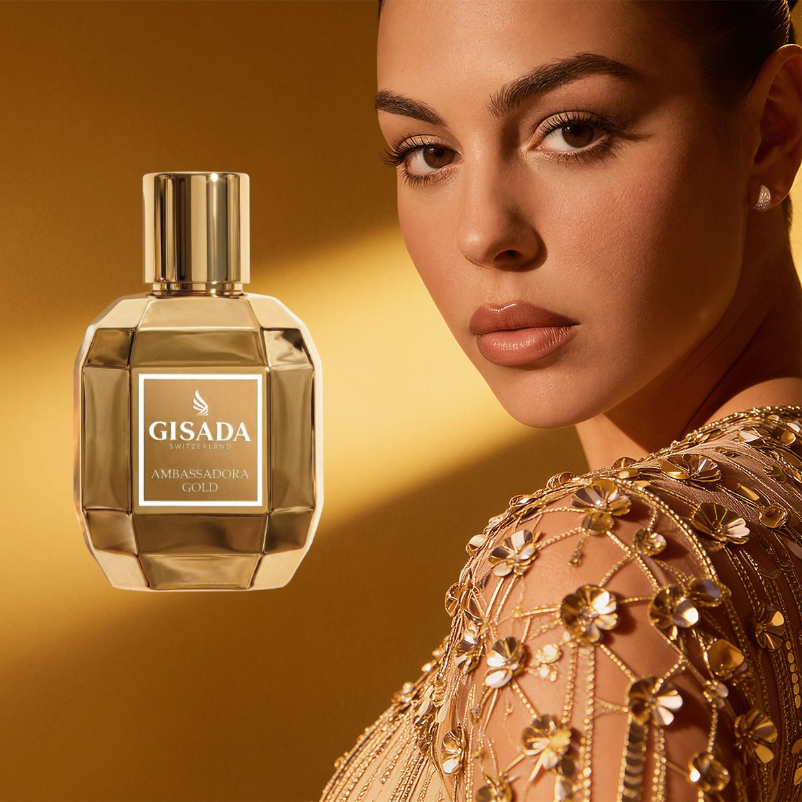 Una donna con un abito dorato posa accanto a Gisada Ambassadora Gold, una fragranza in una bottiglia dorata sfaccettata con un'etichetta bianca.