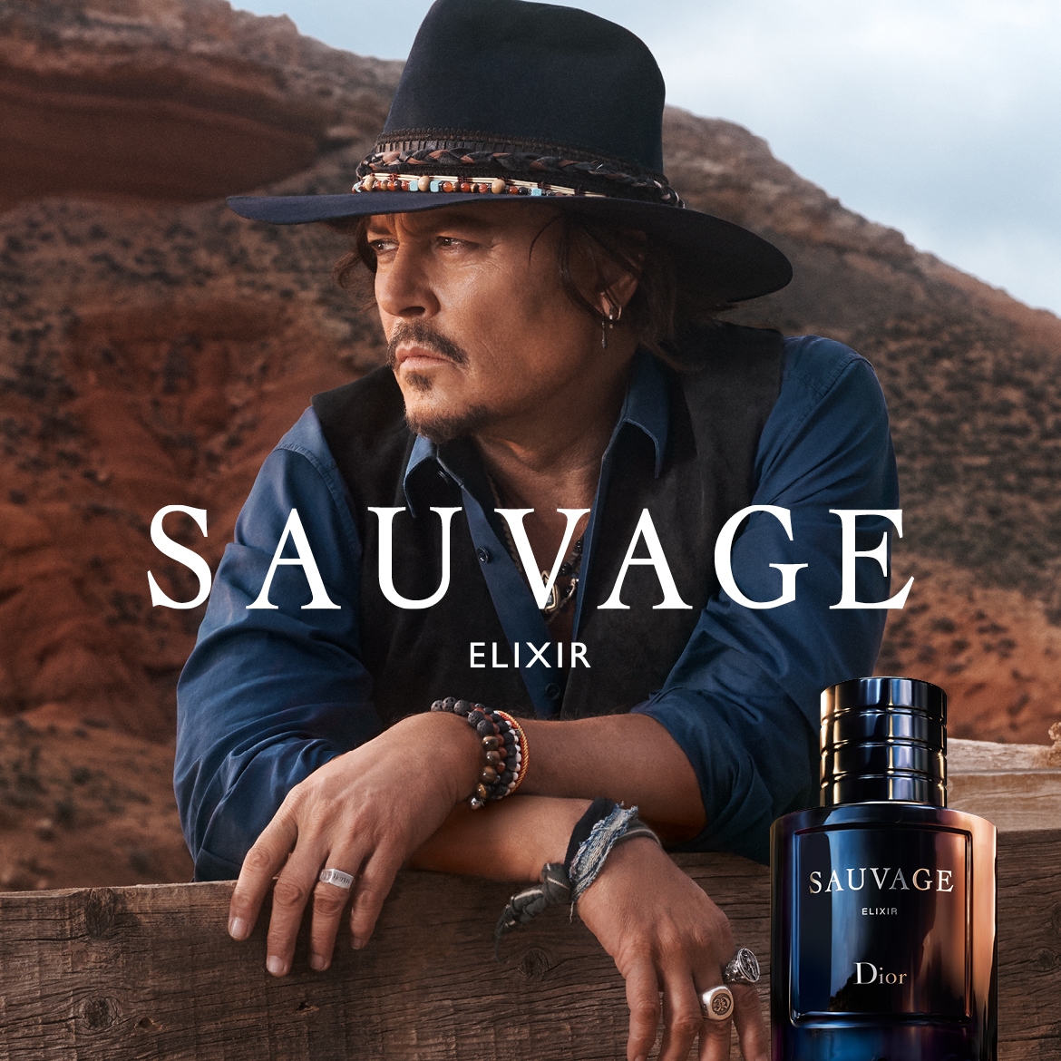 Un uomo con cappello e gilet appoggiato a una recinzione di legno, con una bottiglia di profumo blu scuro Sauvage Elixir di Dior in primo piano.