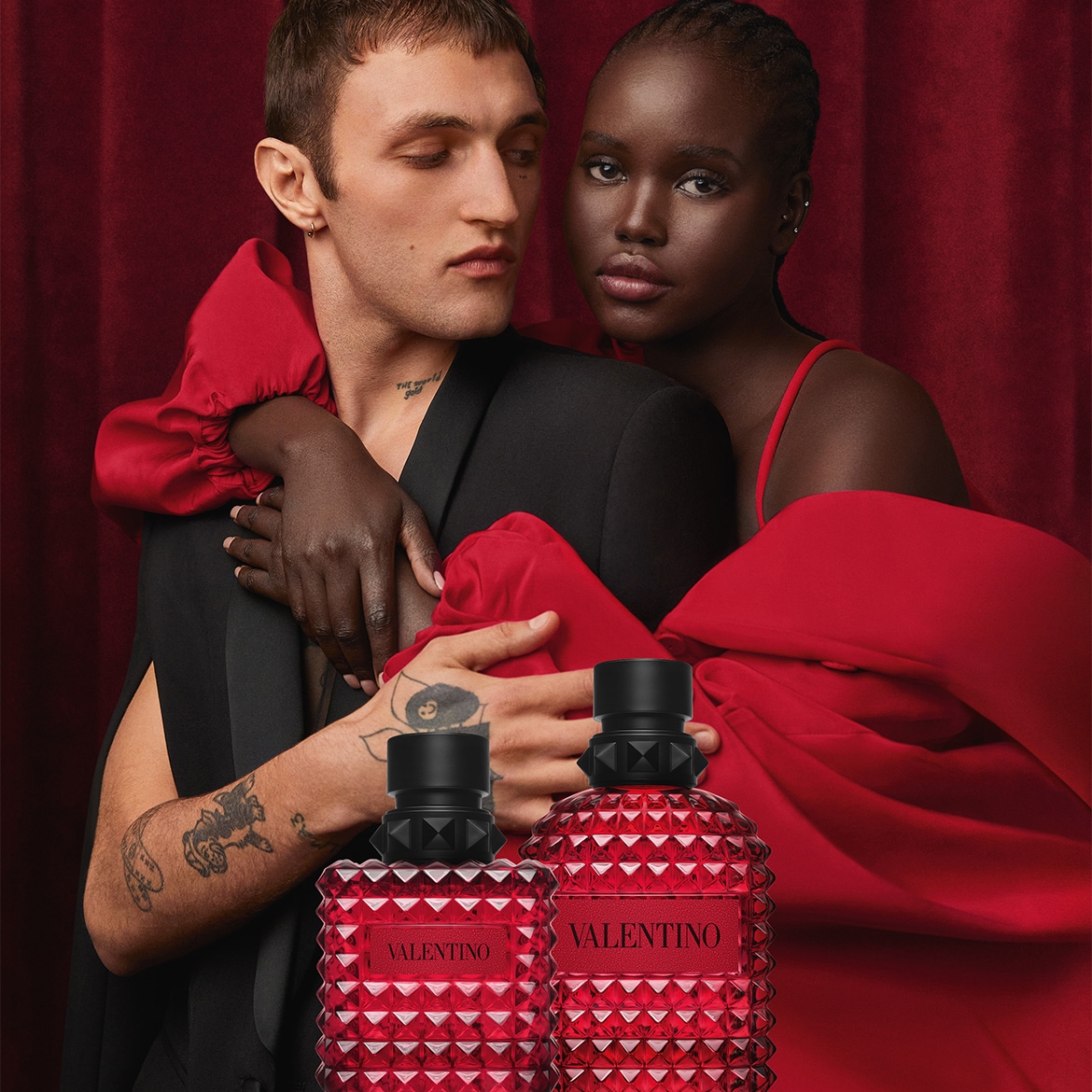 Due modelli, un uomo e una donna, posano con due flaconi di profumo Valentino rosso rubino, con tappi neri borchiati, su uno sfondo di tende rosse.