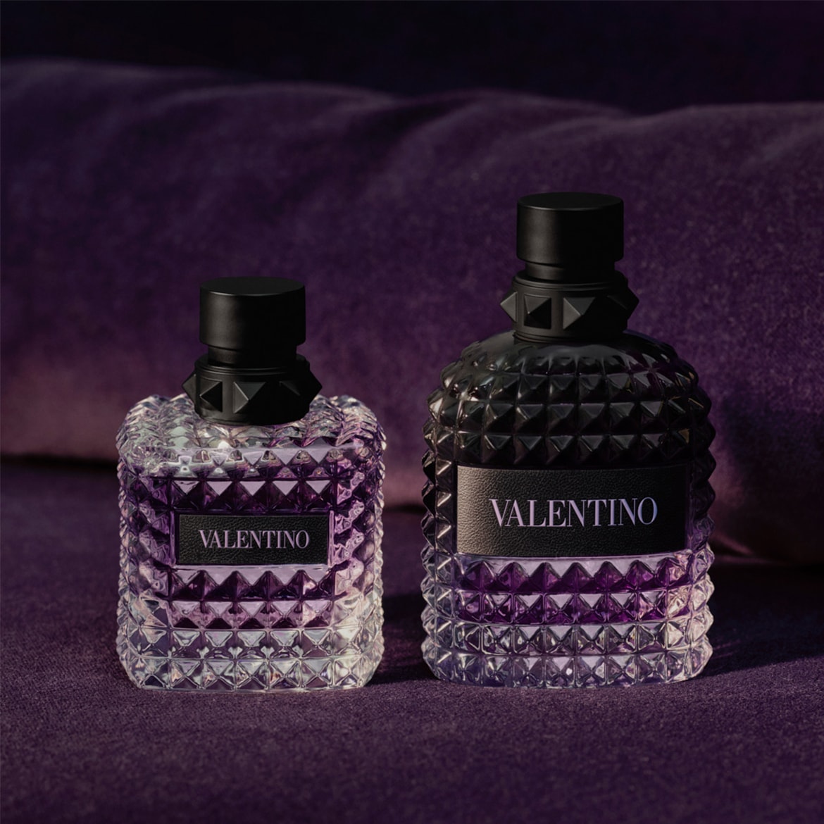 Ecco due flaconi di profumo Valentino su uno sfondo viola scuro, uno chiaro e uno scuro, entrambi con tappi neri borchiati e l'etichetta Valentino.
