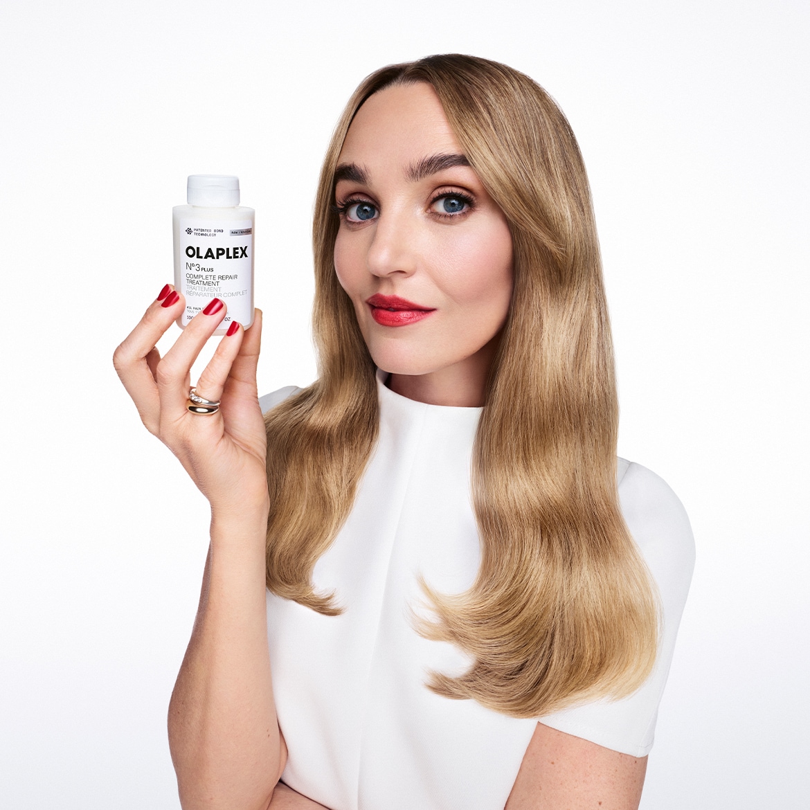 Donna con capelli biondi e rossetto rosso che mostra una bottiglia bianca di Olaplex N.3 Hair Perfector, un trattamento riparatore completo per capelli.