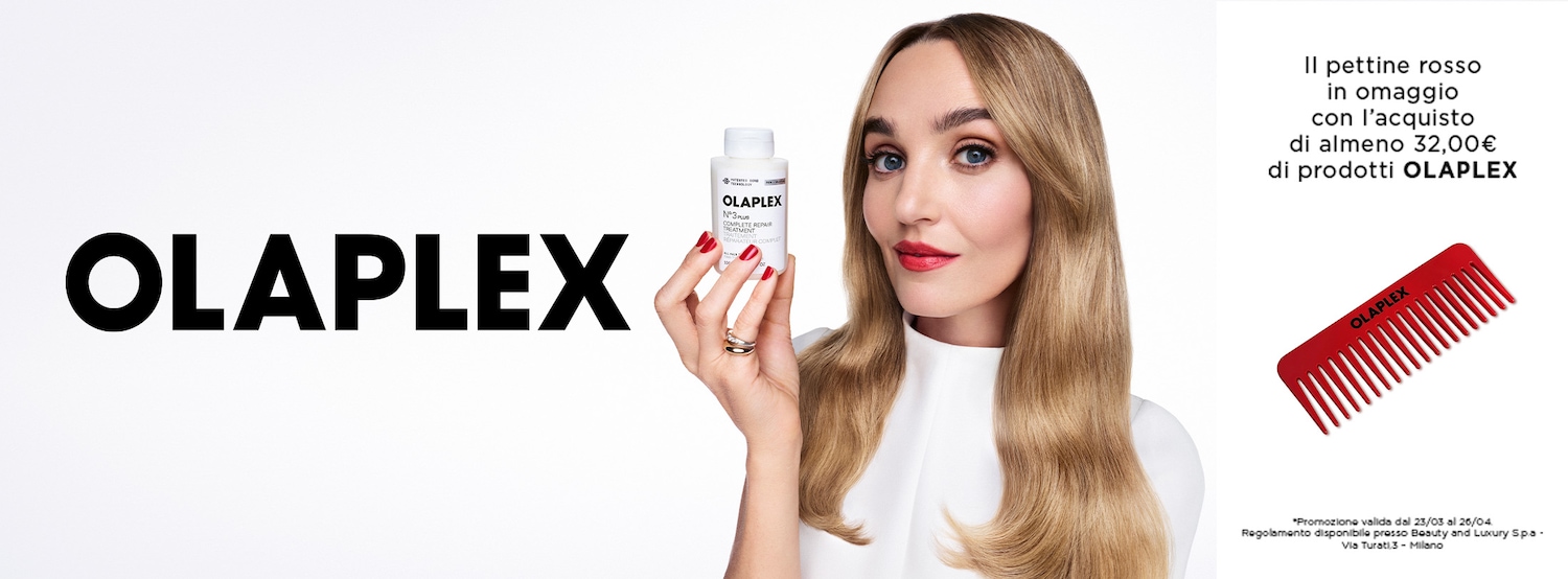 Una donna dai capelli biondi tiene in mano un flacone bianco di Olaplex N°3, mentre a destra si vede un pettine rosso Olaplex in omaggio con l'acquisto di almeno 32,00€ di prodotti Olaplex.