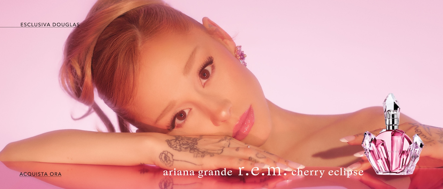 Primo piano di Ariana Grande con un flacone di profumo rosa a forma di fiore, appoggiata su una superficie lucida rosa.