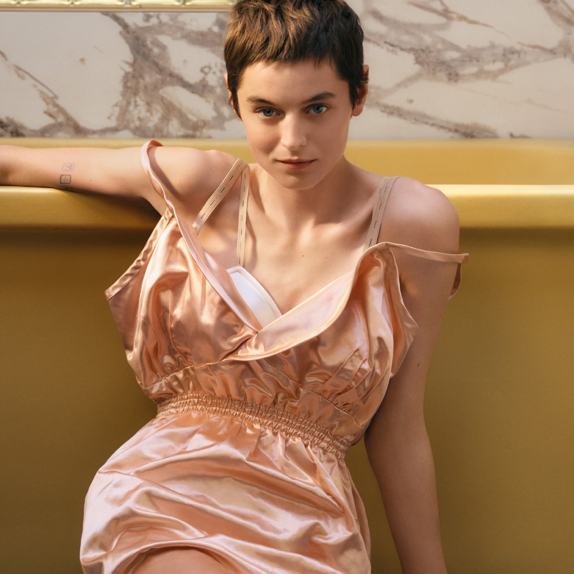 Una donna con i capelli castani corti indossa un abito di seta color pesca, appoggiata a una vasca da bagno gialla, con uno sguardo diretto e sicuro.