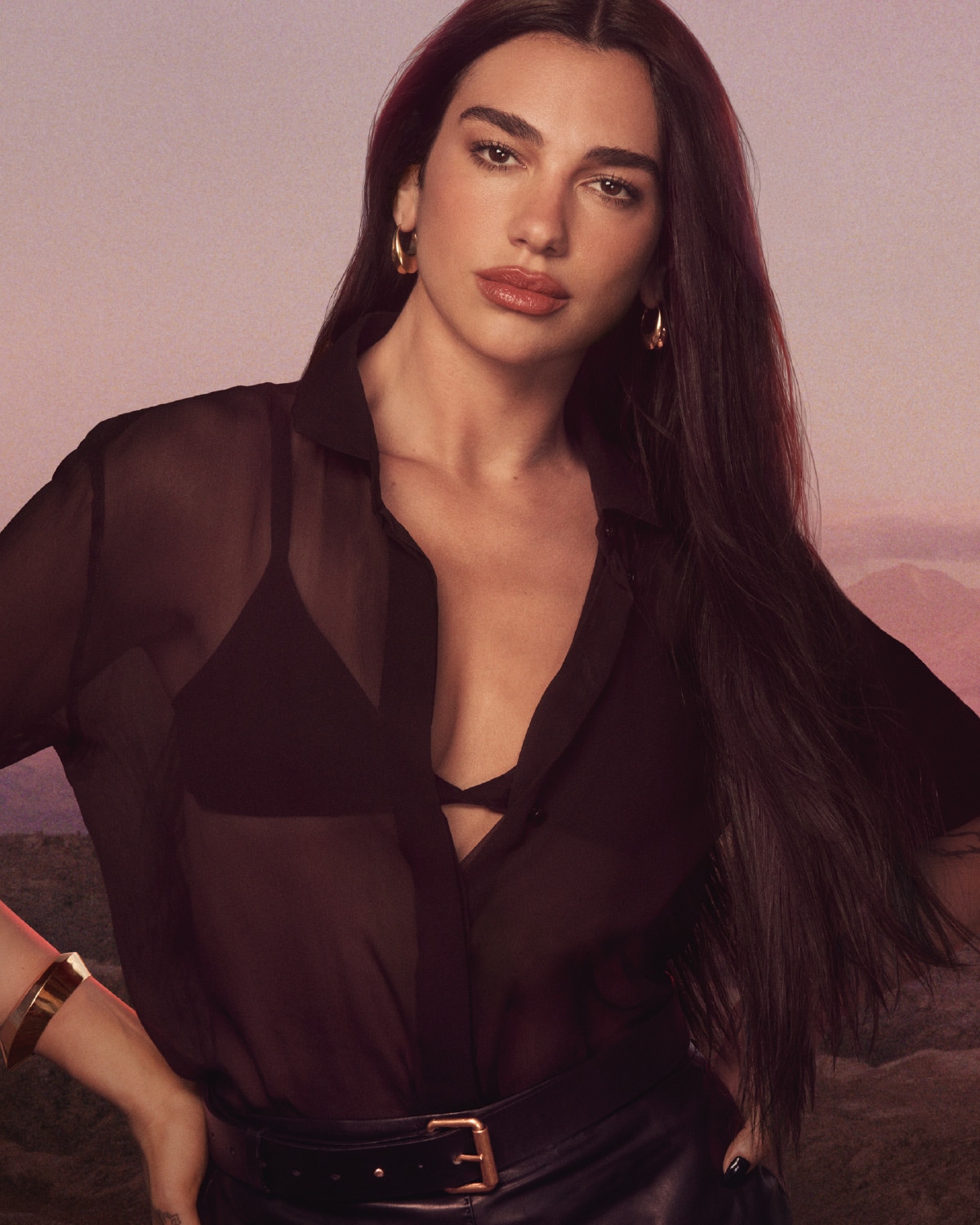 In primo piano, una donna con lunghi capelli scuri indossa una camicia nera trasparente sopra un reggiseno nero, abbinata a una cintura di pelle nera con fibbia dorata e bracciale dorato.