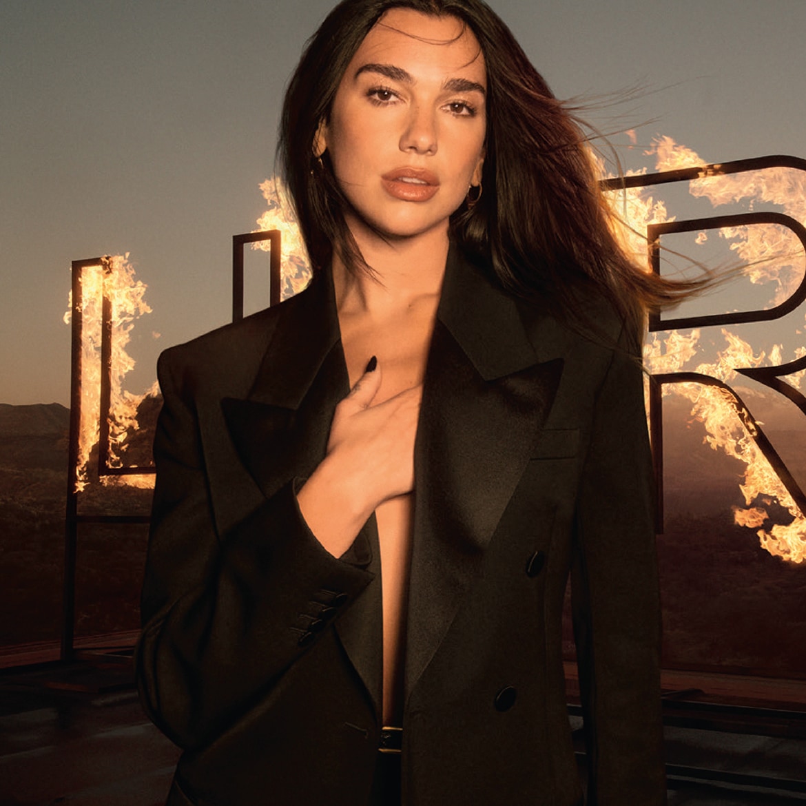 Una donna con blazer nero e capelli castani posa davanti a lettere infuocate, creando un'immagine audace e potente.