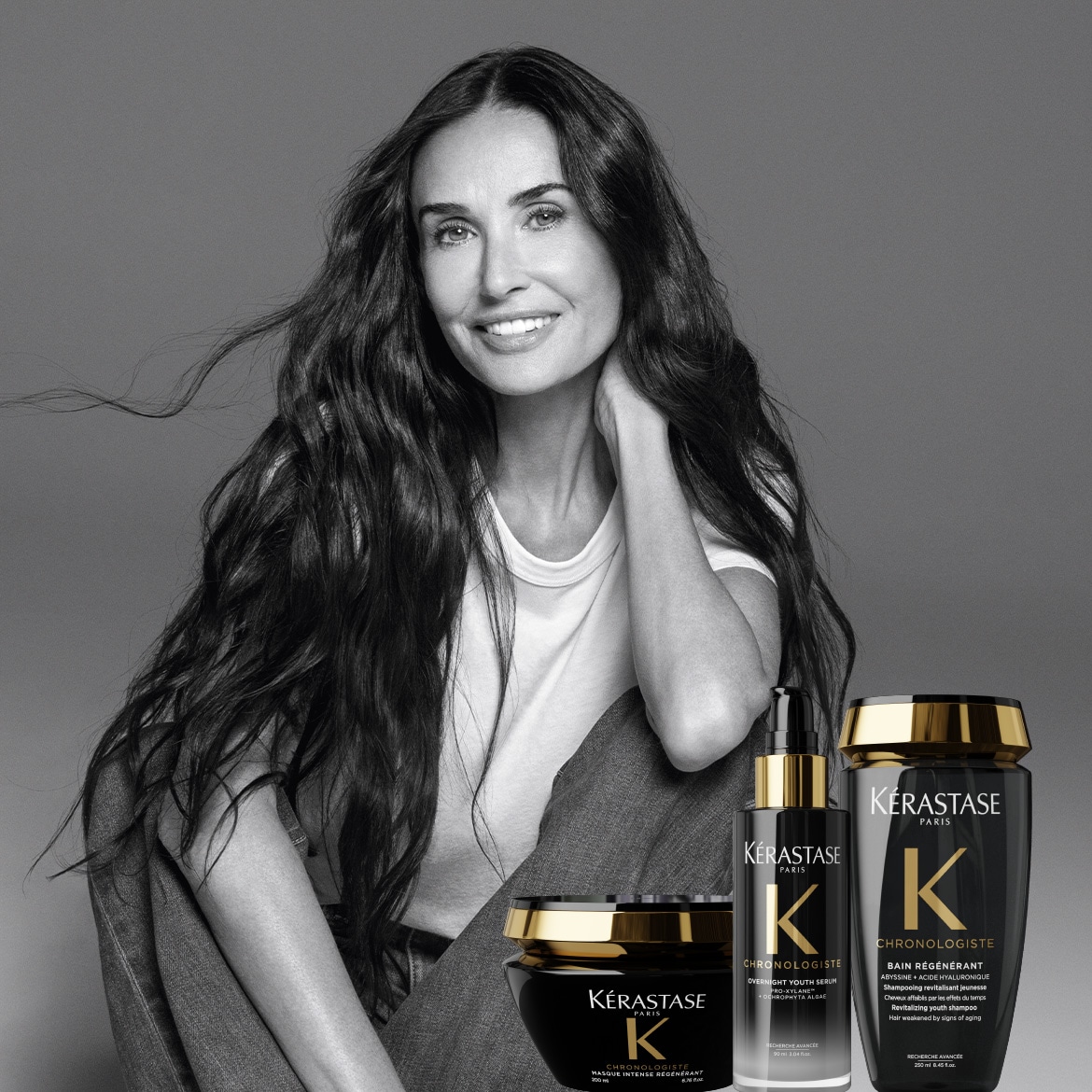 In uno scatto in bianco e nero, Demi Moore è seduta accanto ai prodotti Kérastase Chronologiste, tra cui maschera, siero e shampoo, presentati in confezioni nere e oro.
