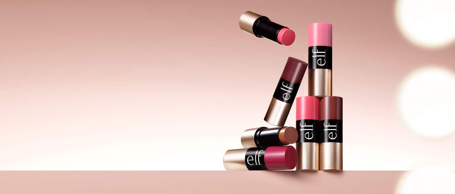 Una collezione di stick per labbra e guance e.l.f. Cosmetics in varie tonalità di rosa, rosso e marrone, con confezione nera e oro, è disposta artisticamente su una superficie rosa chiaro.
