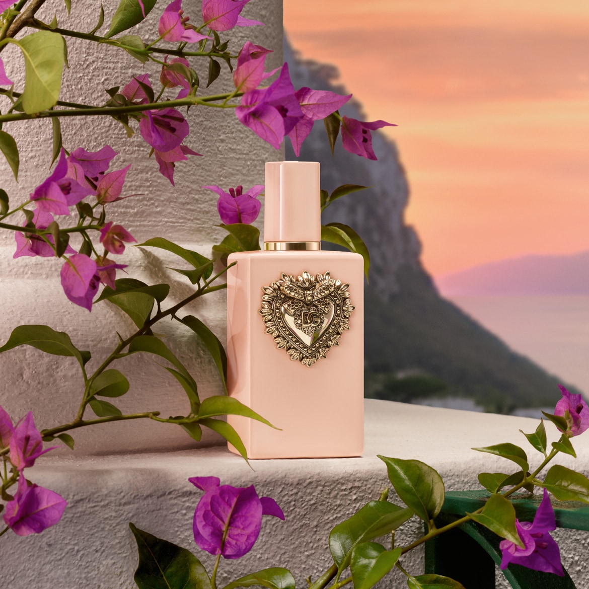 Il profumo Dolce&Gabbana in un flacone rosa chiaro con un cuore dorato, circondato da fiori viola.