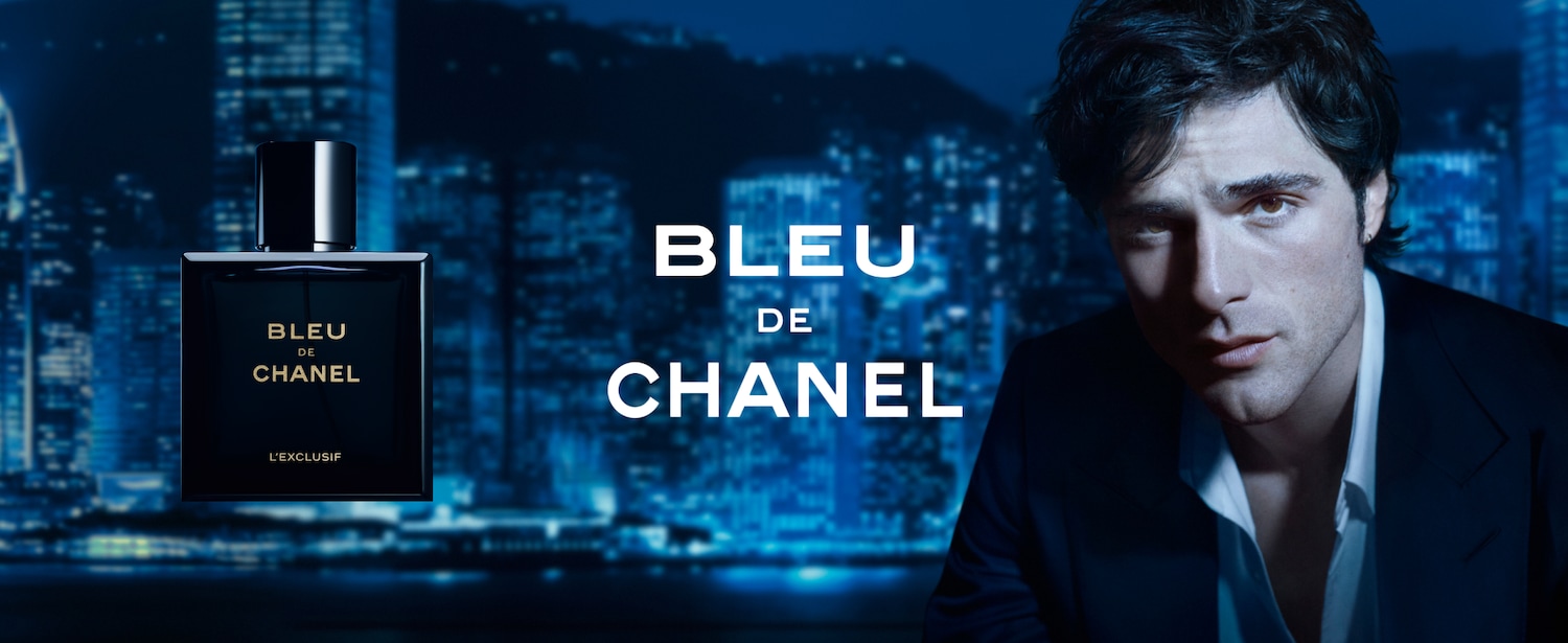 L'immagine mostra una bottiglia di profumo squadrata blu scuro con la scritta dorata "BLEU DE CHANEL" su uno sfondo notturno urbano sfocato e un uomo in giacca.