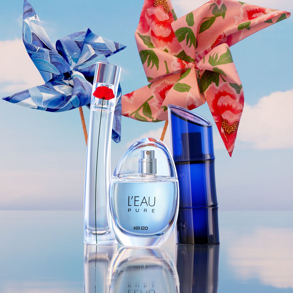 Tre flaconi di profumo Kenzo, uno trasparente con un fiore rosso, uno azzurro a forma di goccia e uno blu scuro, su una superficie riflettente con girandole colorate sullo sfondo.