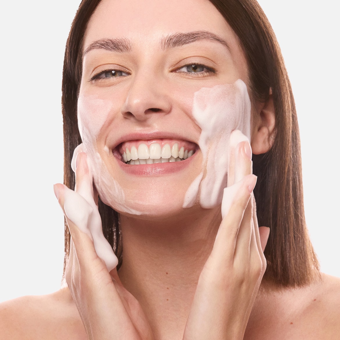 Una donna sorridente si sta lavando il viso con un detergente schiumogeno bianco, massaggiandolo delicatamente sulla pelle.
