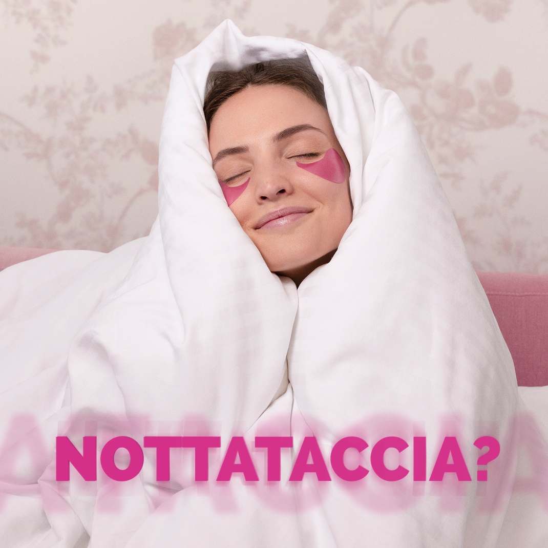 Una donna sorridente è avvolta in un piumone bianco, con patch rosa sotto gli occhi e la scritta "NOTTATACCIA?" in rosa brillante in primo piano.