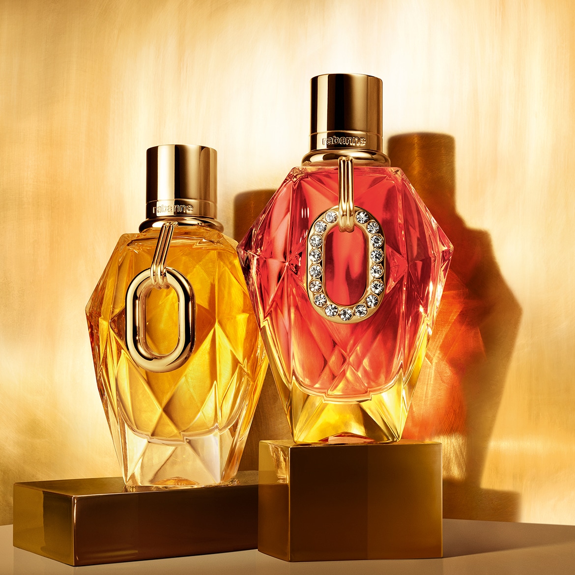 Ecco due flaconi di profumo Rabanne su piedistalli dorati: uno giallo ambrato e l'altro rosso, entrambi con un anello dorato e un tappo cilindrico dorato.