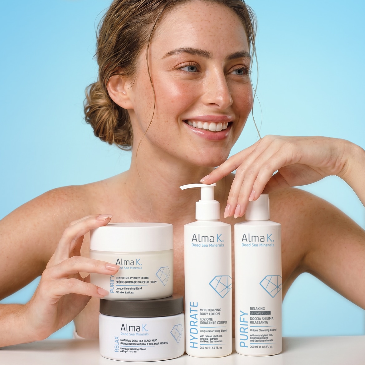 Una donna sorridente mostra i prodotti Alma K. Dead Sea Minerals, tra cui uno scrub corpo delicato, un fango nero naturale, una lozione corpo idratante e un gel doccia rilassante.