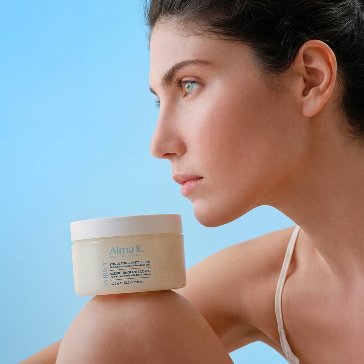 Primo piano di una donna con la pelle chiara e i capelli scuri, che tiene in mano un barattolo bianco di scrub corpo stimolante Alma K. con oli nutrienti e sale del Mar Morto.