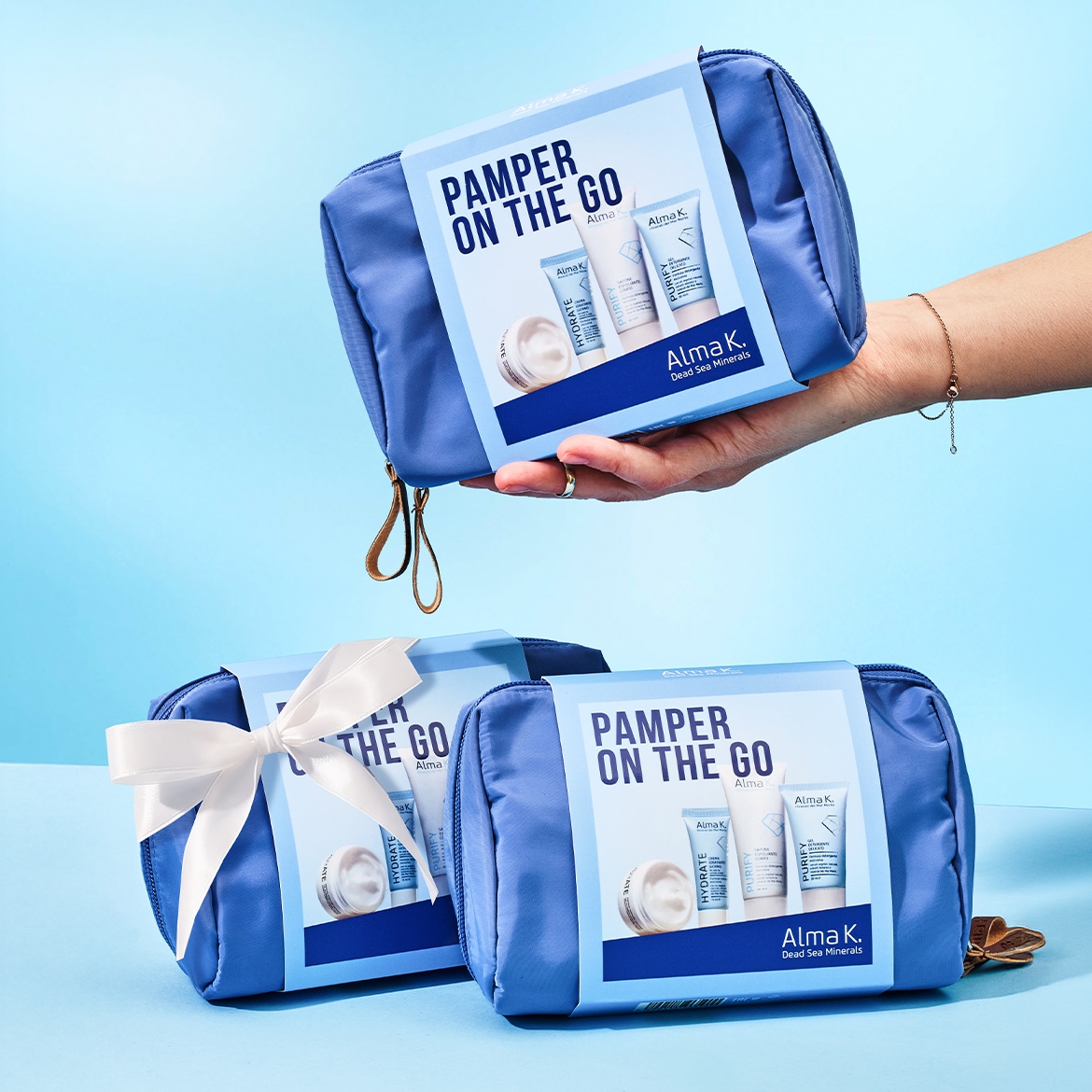 Ecco tre beauty case blu "Pamper On The Go" di Alma K, uno tenuto in mano, gli altri due decorati con un nastro bianco, perfetti per portare sempre con te i tuoi prodotti di bellezza preferiti.