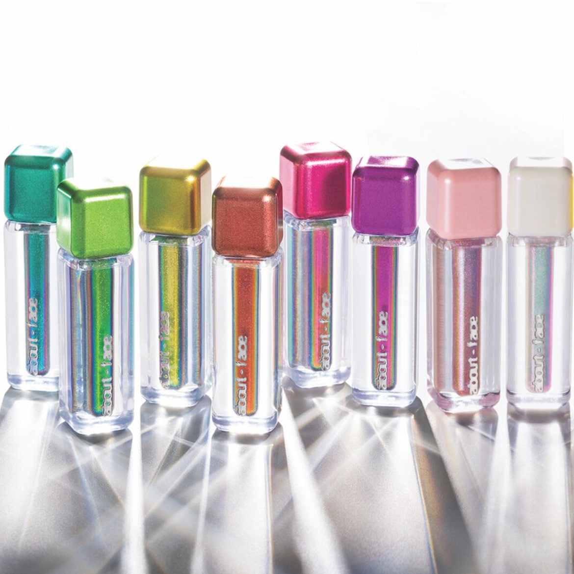 Una fila di sette glitter About-Face in flaconi trasparenti con tappi squadrati colorati, che vanno dal verde al bianco, offrono un arcobaleno di brillantezza.