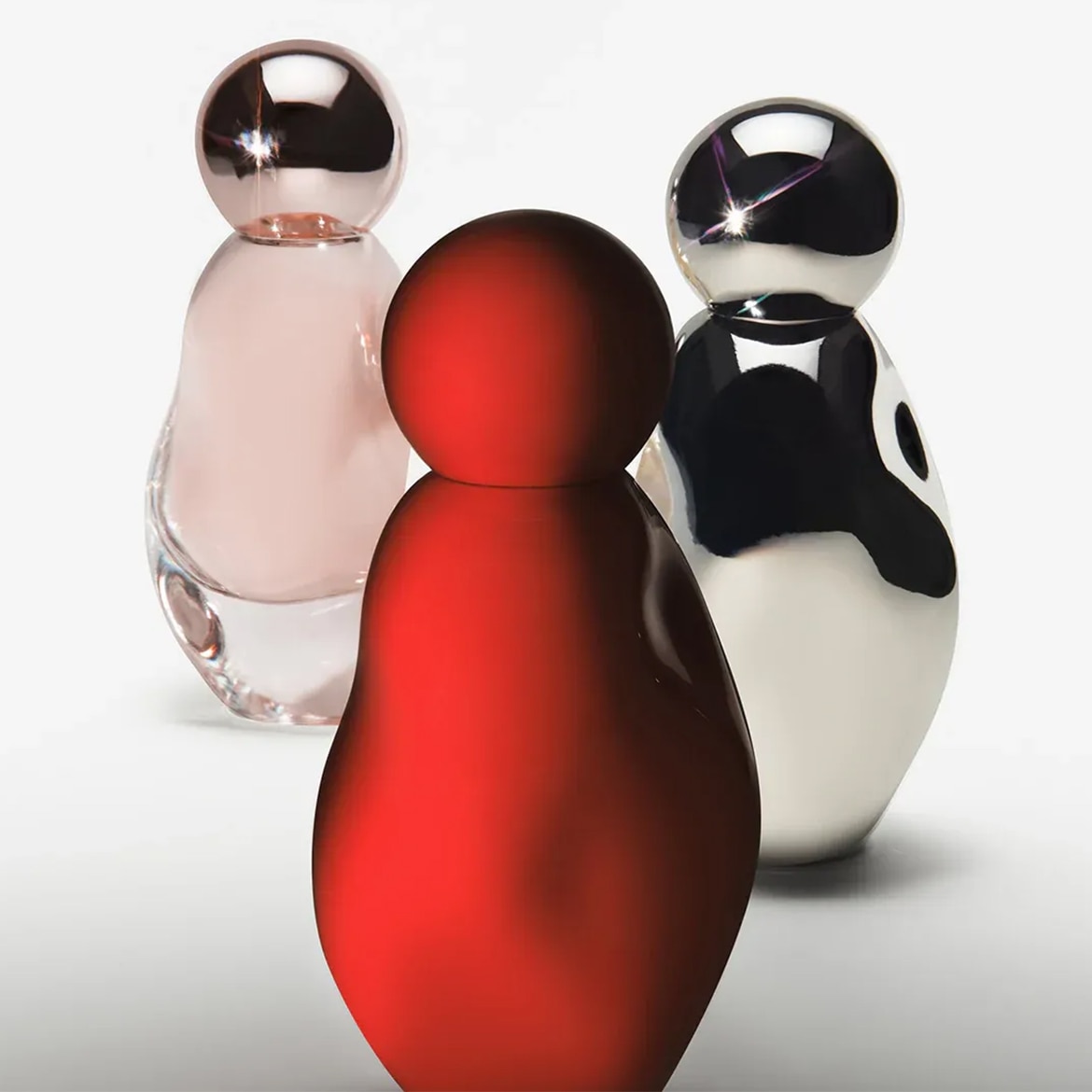 Ecco tre flaconi di profumo colorati a forma di bambola russa: uno rosso acceso, uno rosa pallido con tappo color rame e uno argento e nero lucido.