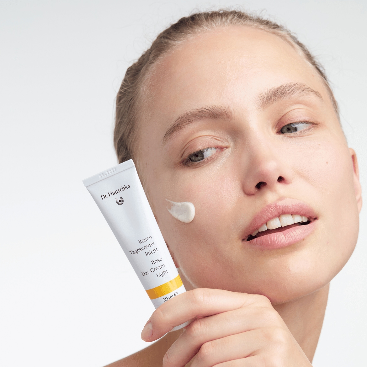 Una donna applica delicatamente la crema da giorno Rose Light di Dr. Hauschka, contenuta in un tubo bianco con dettagli giallo chiaro, per una pelle radiosa e idratata.