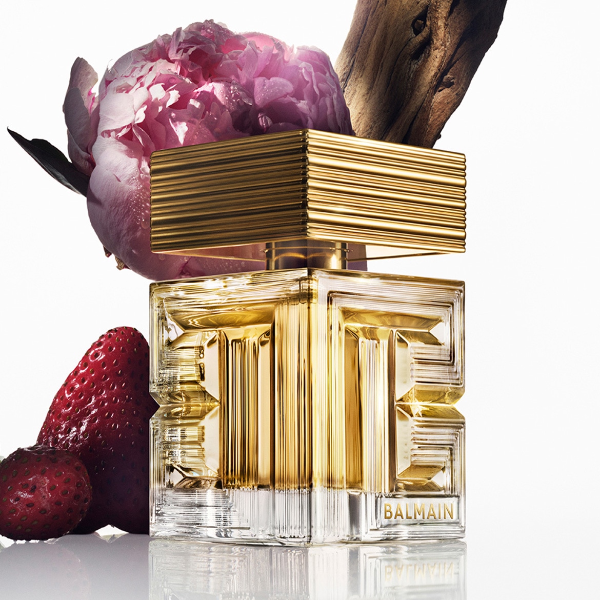 Ecco il profumo Balmain in un flacone di vetro trasparente con tappo dorato, adagiato accanto a una peonia rosa e fragole mature.