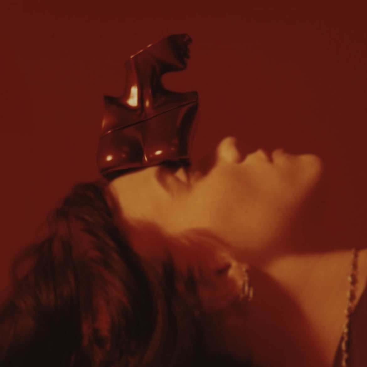 Una donna dai capelli castani è sdraiata con un flacone di profumo rosso a forma di busto femminile in equilibrio sulla fronte.