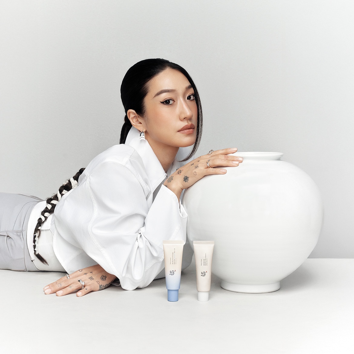 Una donna asiatica con una lunga treccia e una camicia bianca posa accanto a un vaso bianco con due tubi di crema per la pelle, uno con un accento blu e l'altro beige.