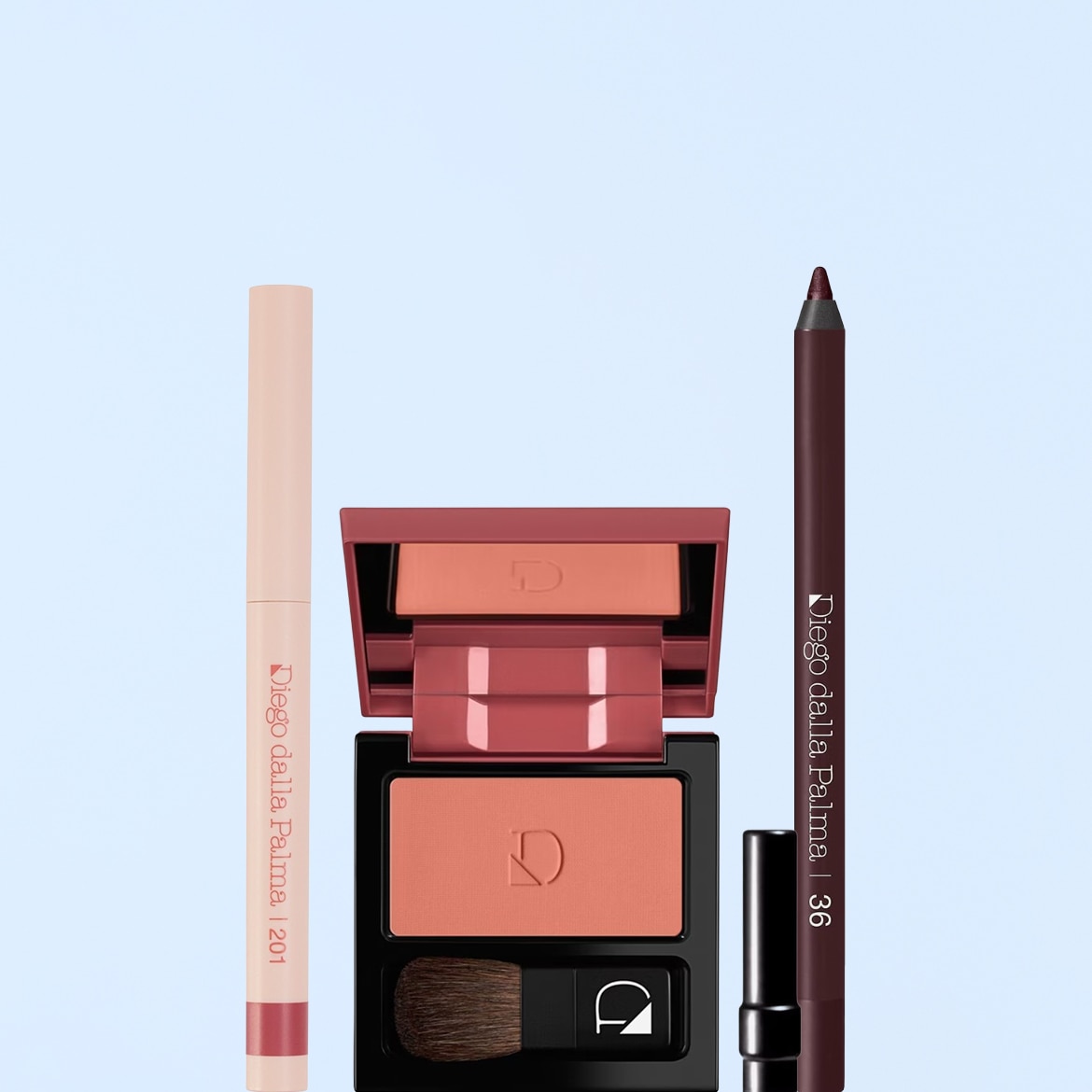 Un set di prodotti Diego dalla Palma, con una matita labbra rosa chiaro, un blush compatto color pesca con pennello e uno specchio, e una matita occhi marrone scuro.