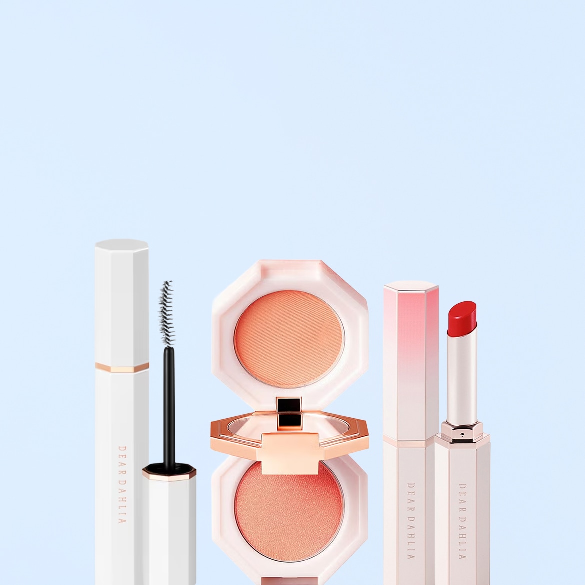 Un mascara bianco con scritte nere, un blush color pesca in una confezione ottagonale aperta e un rossetto rosso in un elegante astuccio bianco e rosa, tutti del marchio Dear Dahlia, sono esposti su uno sfondo azzurro.