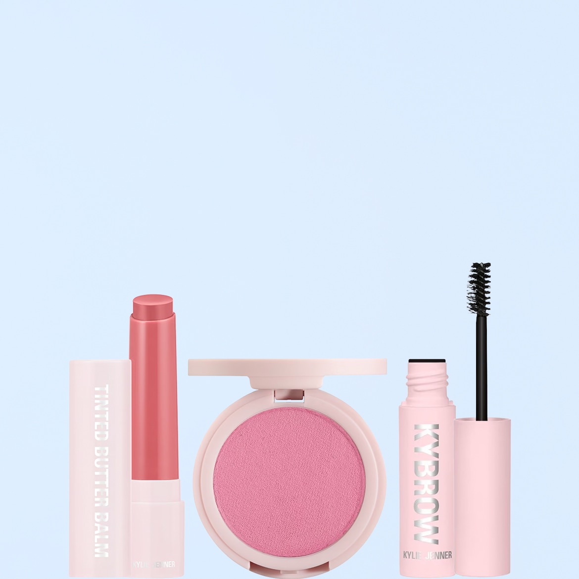 Un set di prodotti Kylie Jenner, tra cui un balsamo labbra colorato rosa chiaro, un fard rosa in una confezione rotonda e un mascara per sopracciglia rosa con la scritta "KYBROW" in verticale.