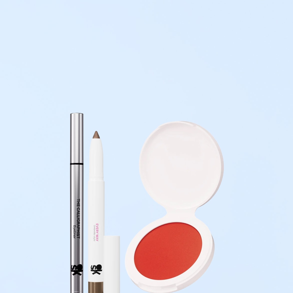 Un eyeliner argentato, una matita marrone e un fard arancione in una confezione bianca aperta sono disposti su uno sfondo azzurro.