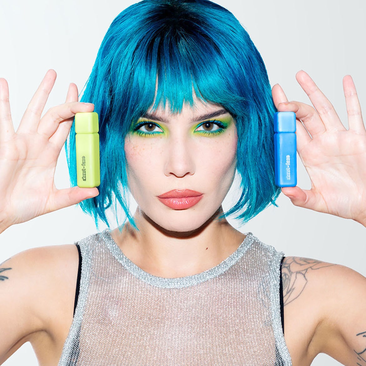 Una modella dai capelli blu elettrico e ombretto verde lime tiene in mano due flaconi di trucco about-face, uno verde chiaro e uno blu.