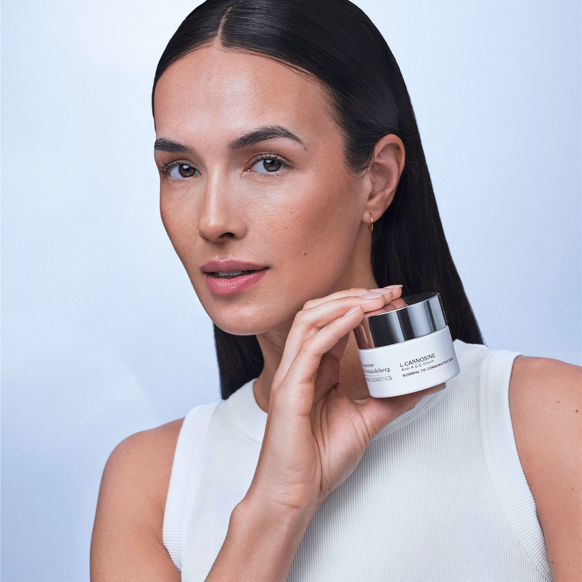 Donna con lunghi capelli scuri tiene in mano un vasetto bianco di crema L-CARNOSINE Anti-A.G.E. Cream per pelli normali o miste di Susanne Kaufmann.