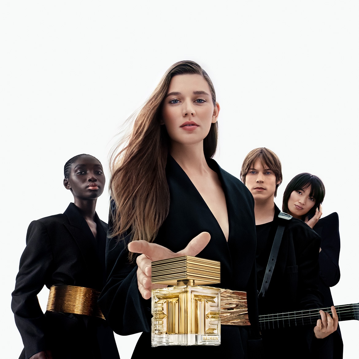 Una donna con lunghi capelli castani tiene in mano una bottiglia di profumo dorata Balmain, mentre dietro di lei ci sono tre persone vestite di nero, creando un'atmosfera elegante e misteriosa.