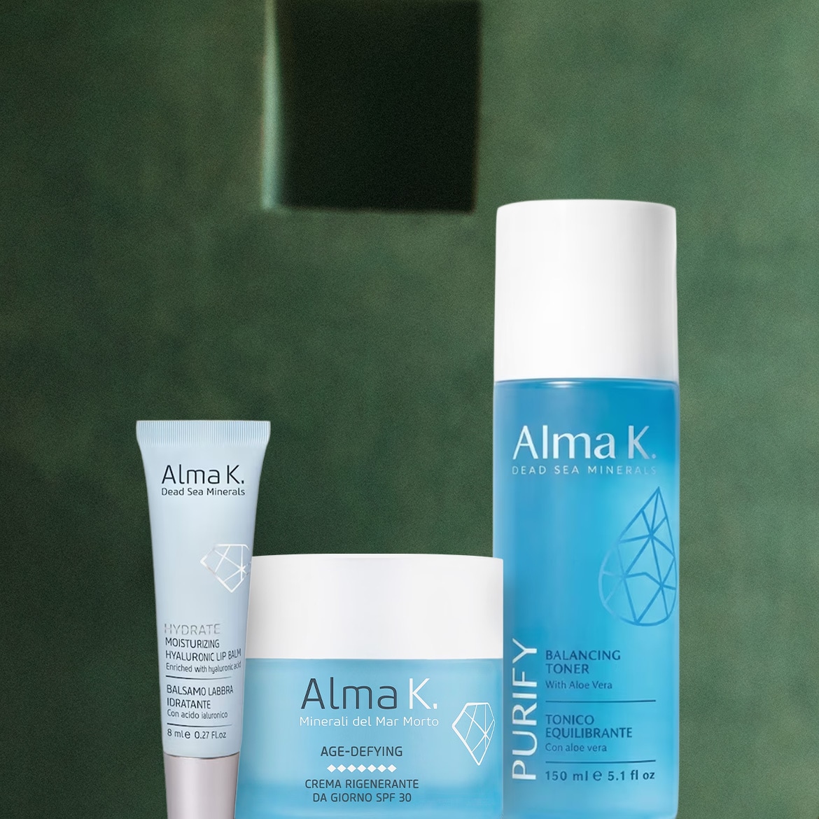 Tre prodotti Alma K. Dead Sea Minerals sono esposti su uno sfondo verde scuro: un balsamo labbra idratante, una crema rigenerante giorno con SPF 30 e un tonico equilibrante con aloe vera.
