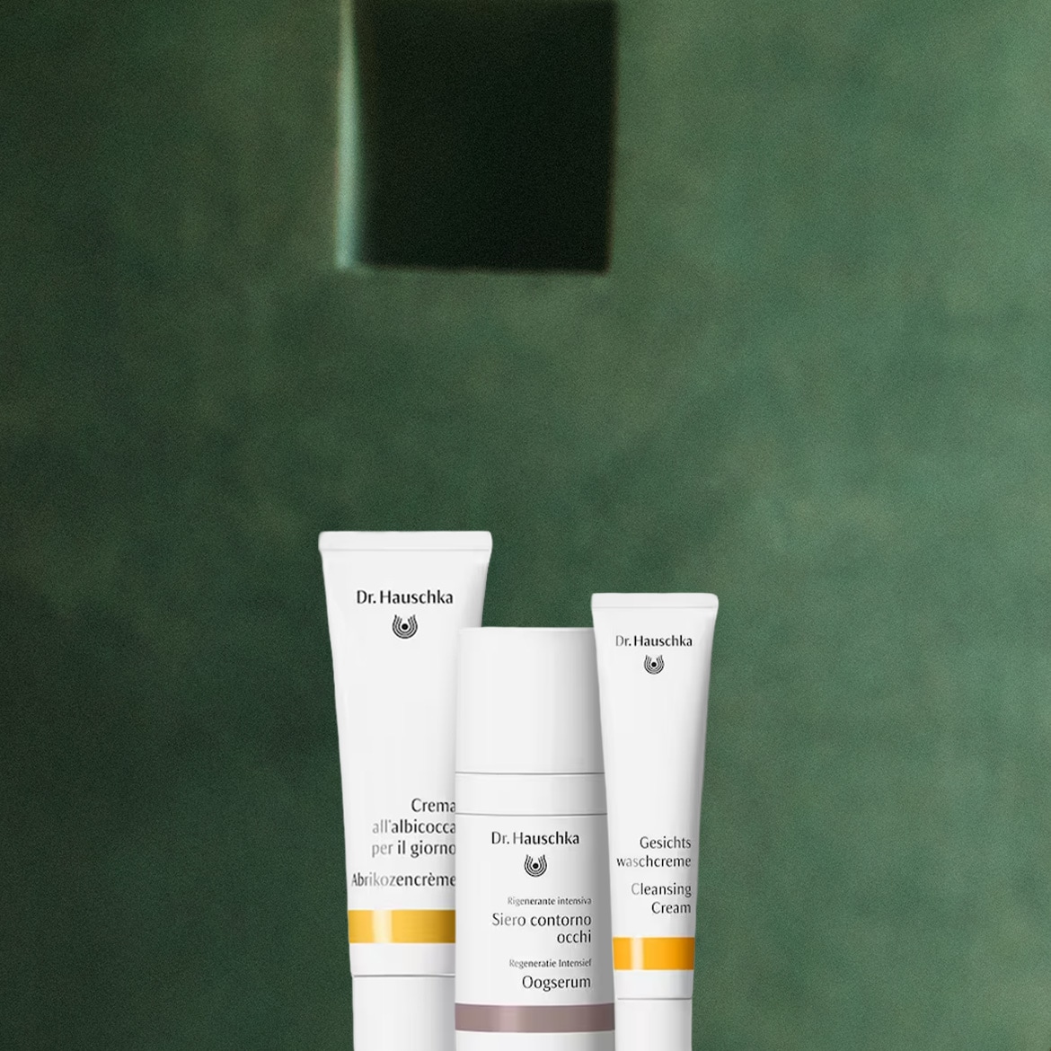 Tre prodotti Dr. Hauschka, tra cui una crema viso all'albicocca, un siero contorno occhi e una crema detergente, sono disposti su uno sfondo verde scuro.