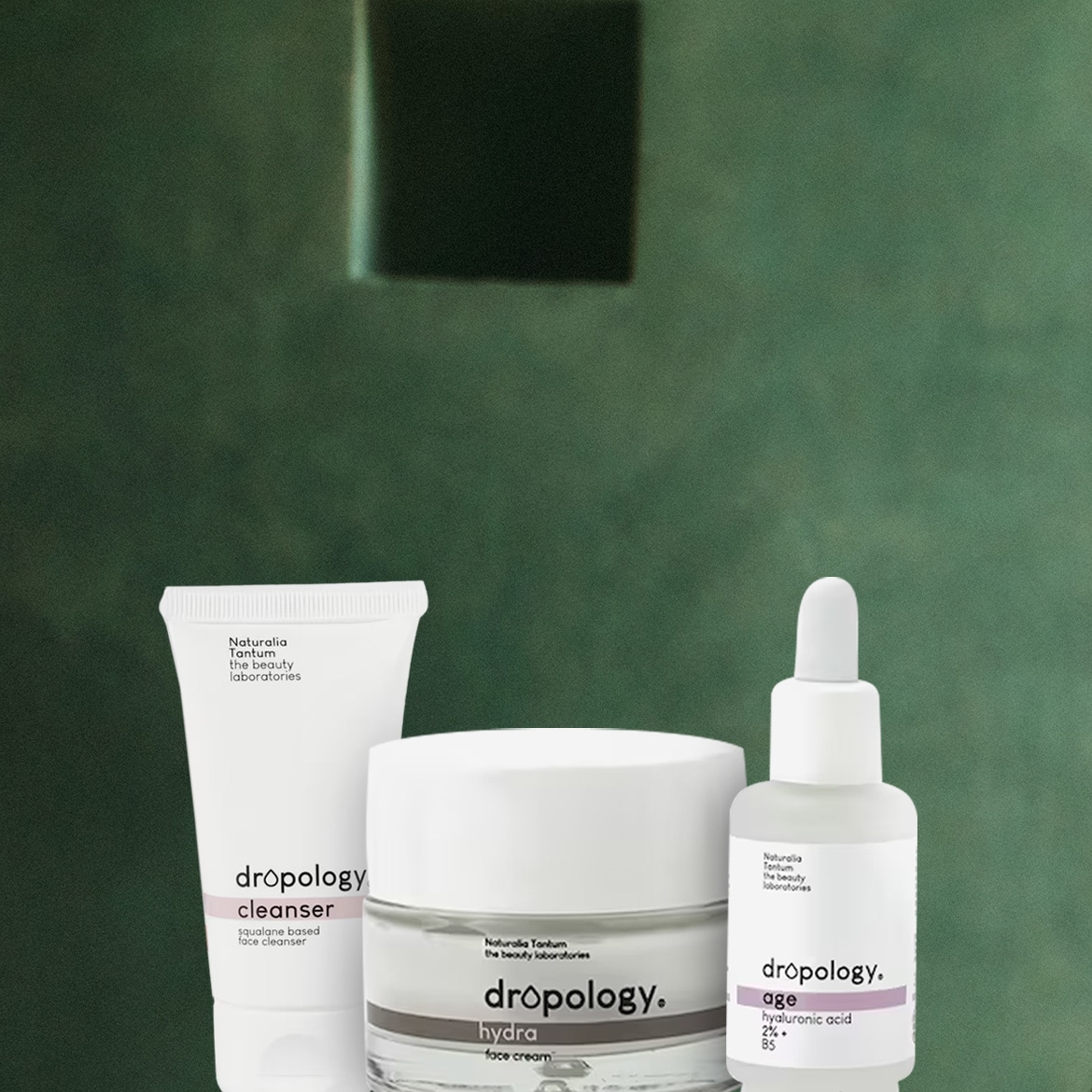 Tre prodotti Dropology di Naturalia Tantum, tra cui un detergente viso a base di squalano, una crema viso idratante e un siero viso anti-età con acido ialuronico al 2%, sono esposti su uno sfondo verde scuro.