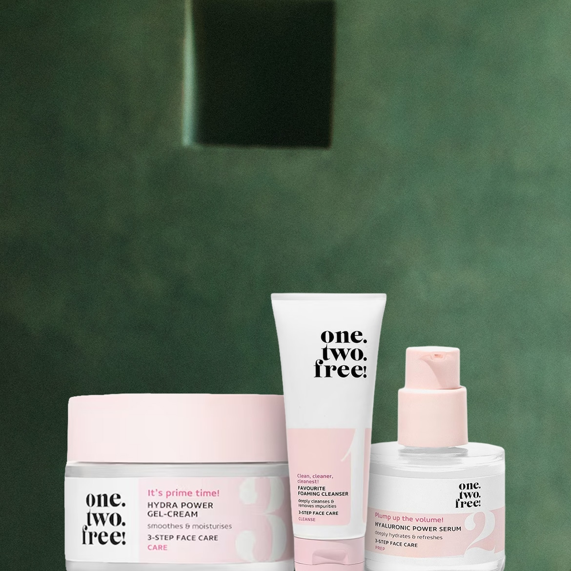 Tre prodotti One.Two.Free! sono esposti su uno sfondo verde scuro: una crema gel Hydra Power in un vasetto rosa chiaro, un detergente schiumogeno in un tubetto bianco e un siero Hyaluronic Power in una bottiglia trasparente con erogatore rosa.