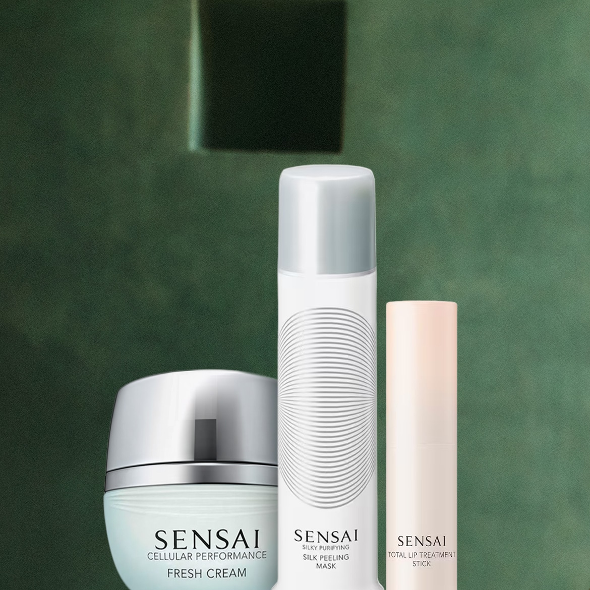 Tre prodotti Sensai sono esposti su uno sfondo verde scuro: una crema idratante in un vasetto azzurro con tappo argentato, una maschera peeling in un flacone bianco con tappo argentato e uno stick per labbra color pesca.