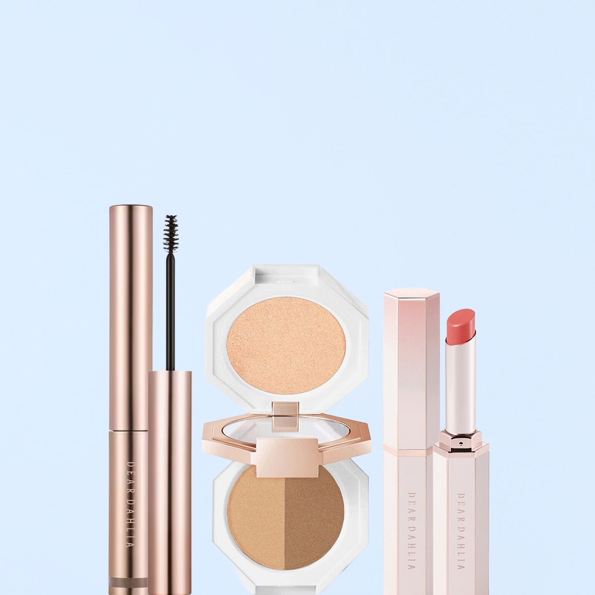 Un mascara color oro rosa con scovolino nero, una palette di ombretti bianca con due tonalità di marrone e un rossetto rosa chiaro in un elegante packaging esagonale.