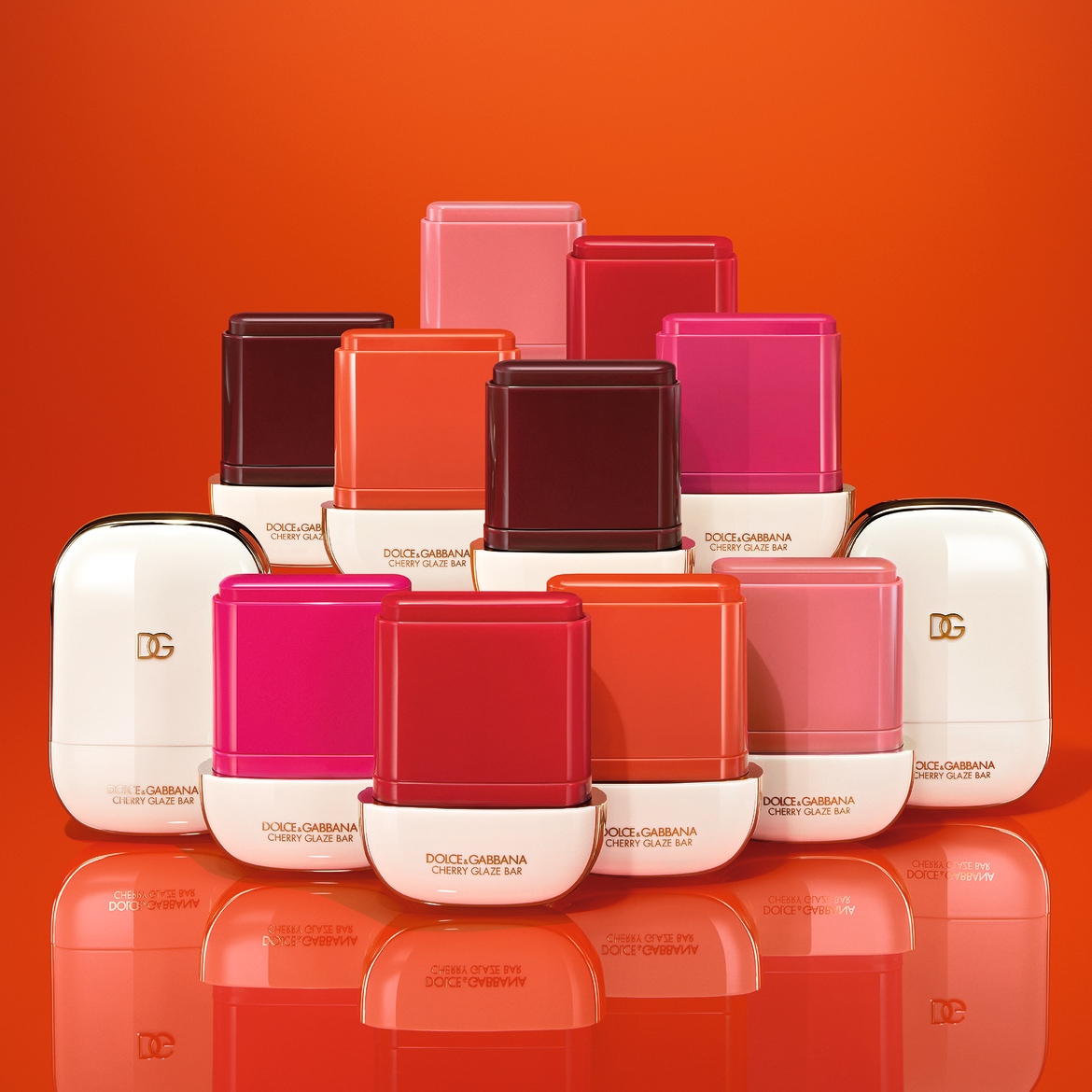 Ecco una collezione di lucidalabbra Dolce&Gabbana Cherry Glaze Bar in diverse tonalità di rosso, rosa e marrone, presentati su uno sfondo arancione lucido.