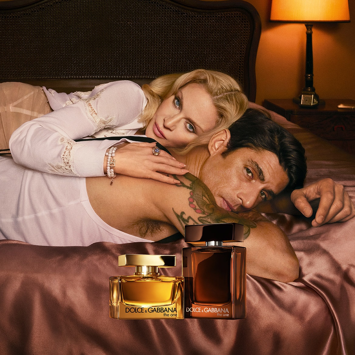 Una coppia distesa su un letto color bronzo con due flaconi di profumo Dolce & Gabbana "The One", uno dorato e uno marrone scuro, in primo piano.