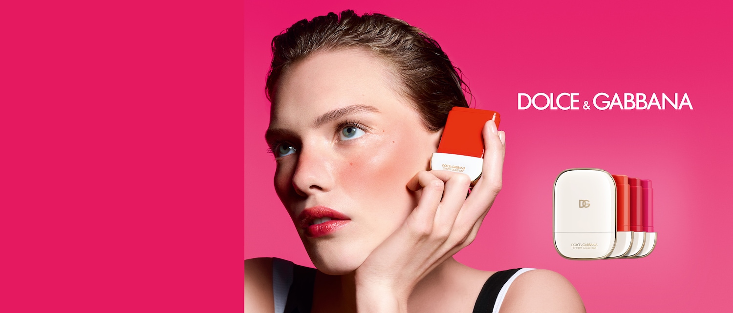 Una modella con la pelle luminosa tiene in mano un Cherry Glaze Bar di Dolce&Gabbana color corallo, con il logo del marchio in evidenza su uno sfondo fucsia.