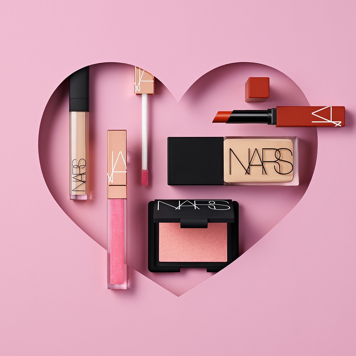 Una selezione di cosmetici NARS, tra cui un correttore, un lucidalabbra rosa scintillante, un blush pesca, una cipria compatta e un rossetto rosso-arancio, disposti all'interno di una forma di cuore rosa.