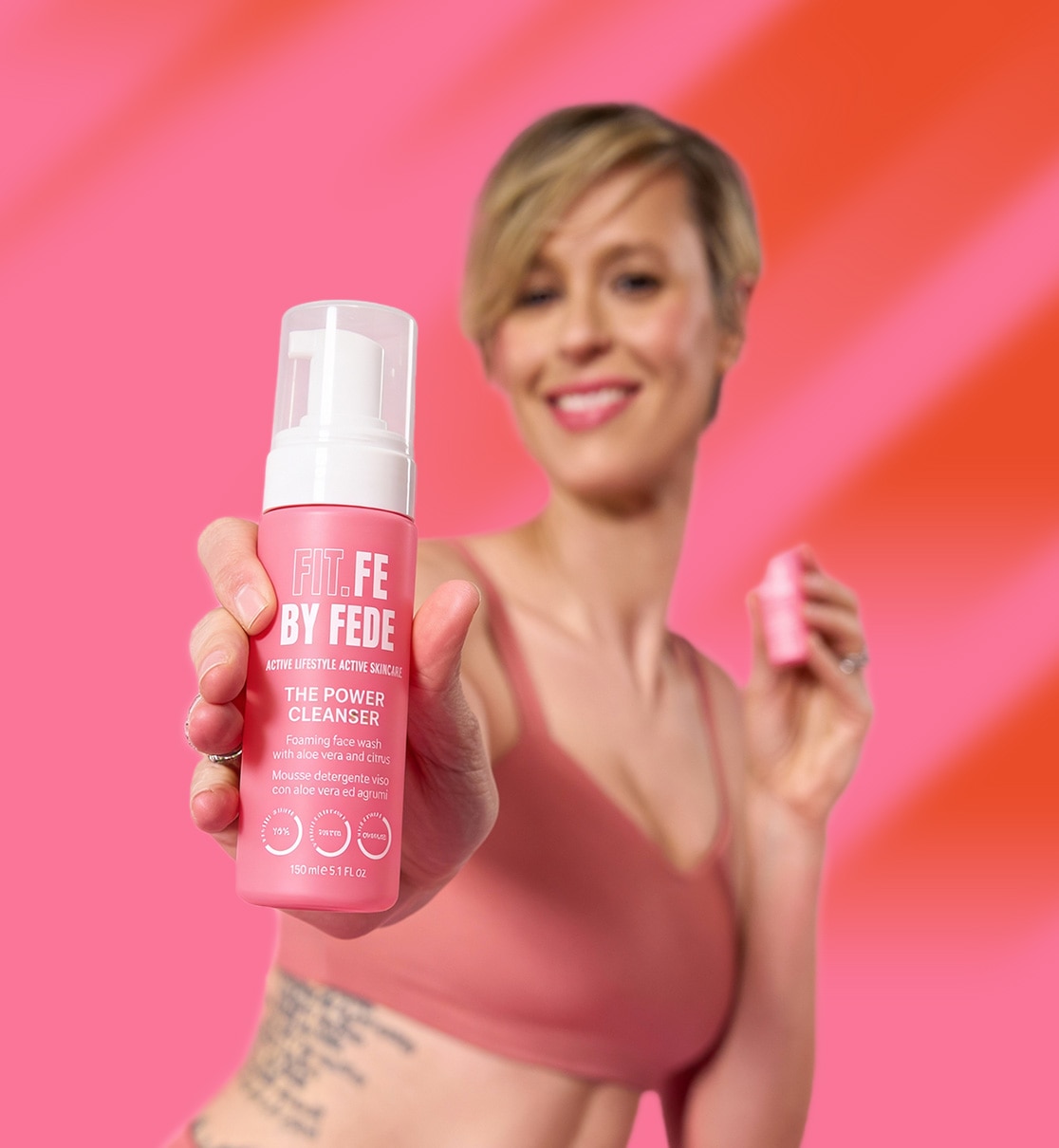 Una donna sorridente mostra il detergente viso FIT.FE BY FEDE in un flacone rosa con scritte bianche, ideale per la cura della pelle attiva.