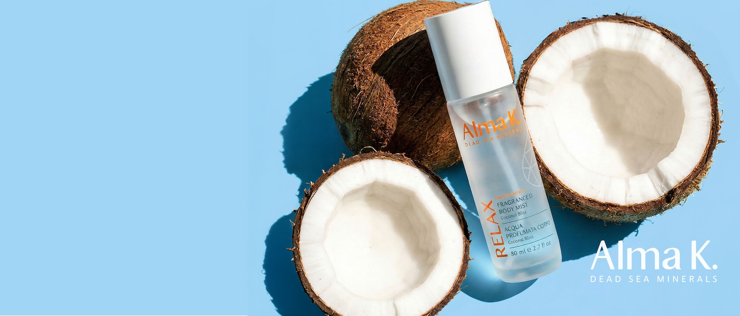 Ecco il body mist profumato Alma K. Dead Sea Minerals, edizione limitata, con aroma di cocco, presentato accanto a noci di cocco su sfondo azzurro.