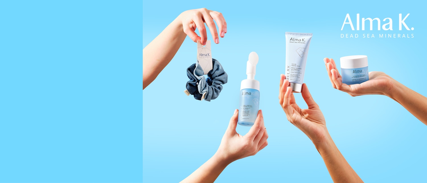 Su sfondo azzurro, mani femminili mostrano prodotti Alma K: uno scrub viso in un tubo bianco-azzurro, una mousse detergente con spazzola e una crema giorno idratante in un vasetto bianco-azzurro, tutti con logo.