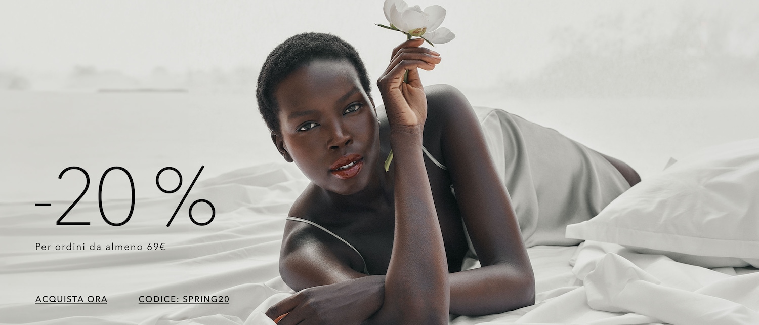 Una donna dai capelli corti e la pelle scura è sdraiata su un letto bianco, con in mano un fiore bianco, e annuncia uno sconto del 20% per ordini superiori a 69€ con il codice SPRING20. Acquista ora!