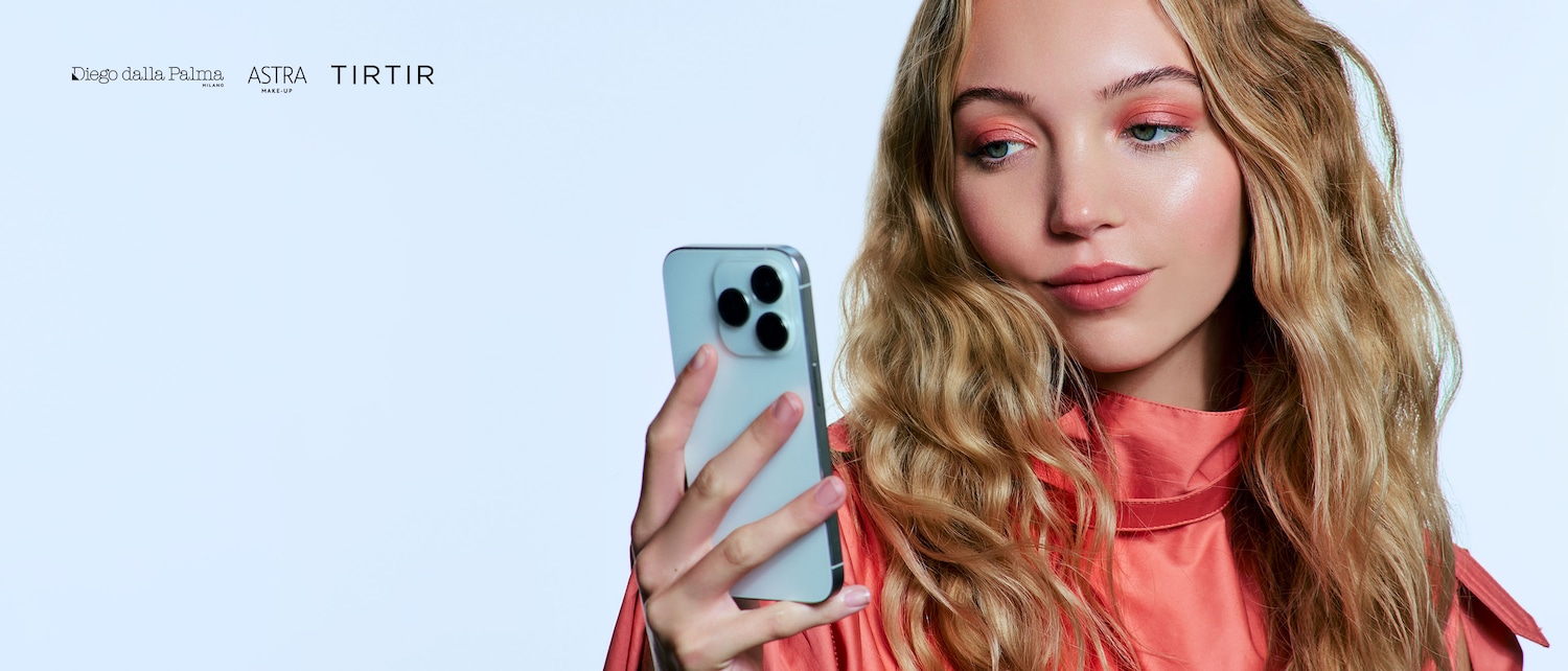 Primo piano di una modella bionda con ombretto pesca e labbra lucide, che tiene in mano un telefono color argento e mostra un look fresco e moderno.
